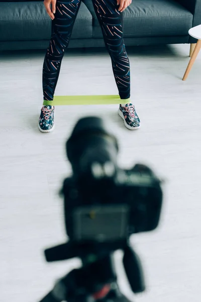 Foco seletivo de esportista com banda de resistência em pernas de pé perto da câmera digital em casa — Fotografia de Stock