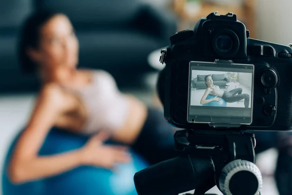 Selective focus of digital camera on tripod near sportswoman training on fitness ball at home — Stock Photo