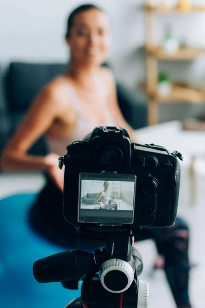 Selective focus of digital camera on tripod near woman in sportswear sitting on fitness ball at home — Stock Photo