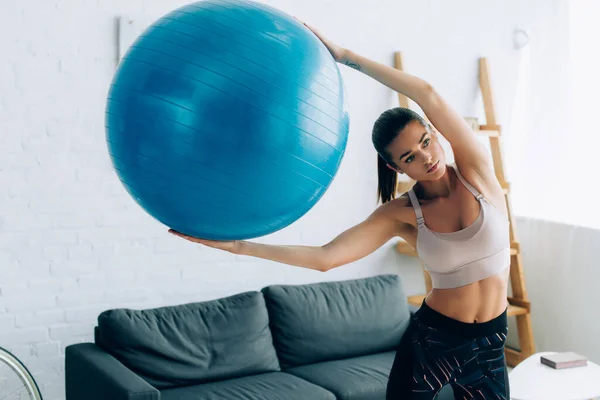 Mujer joven en ropa deportiva sosteniendo la pelota de fitness en la sala de estar - foto de stock