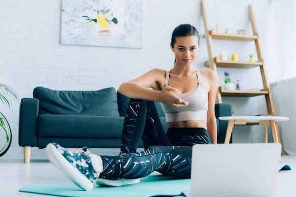 Selective focus of sportswoman pointing with hand at laptop while sitting on fitness mat at home — Stock Photo
