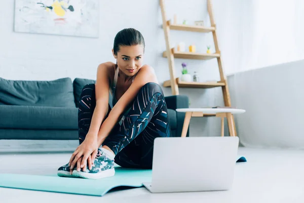 Selective focus of sportswoman sitting on fitness mat near laptop on floor at home — Stock Photo