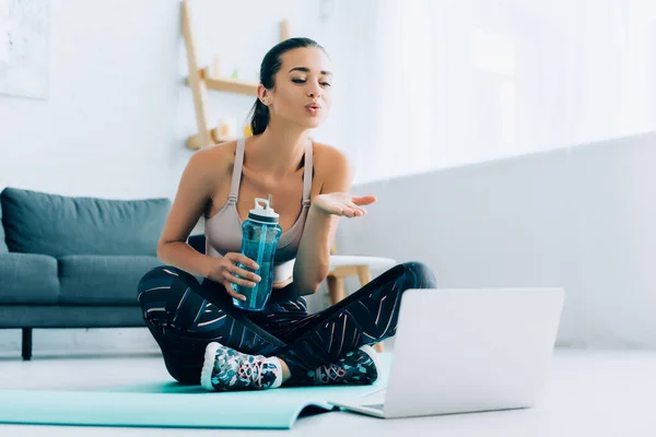 Выборочный фокус молодой спортсменки, дующей воздушным поцелуем в ноутбук, держа спортивную бутылку на коврике для фитнеса — стоковое фото