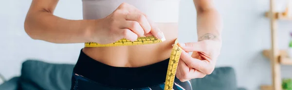 Panoramic crop of fit sportswoman measuring waist at home — Stock Photo