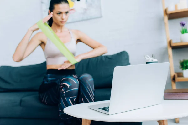 Selective focus of laptop on coffee table near sportswoman using resistance band on couch — Stock Photo