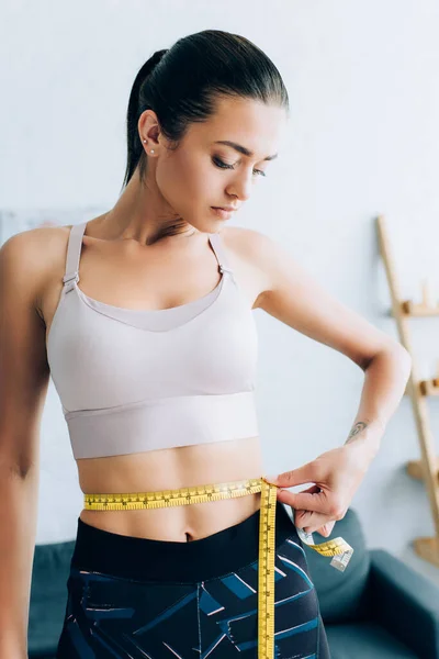 Brunette sportswoman measuring waist with tape in living room — Stock Photo