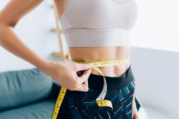 Cropped view of woman in sportswear measuring waist with tape at home — Stock Photo