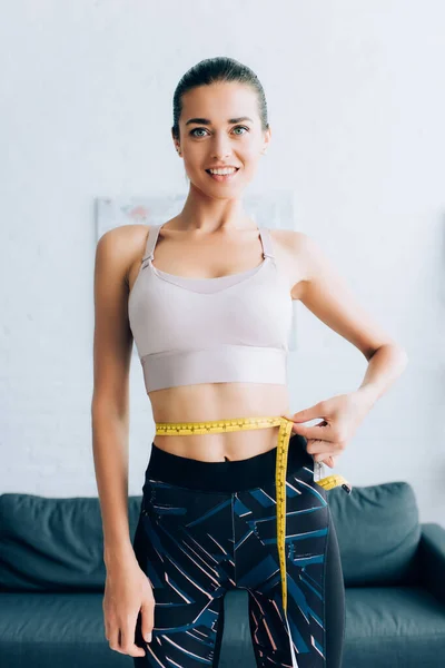 Young sportswoman looking at camera while measuring waist with tape at home — Stock Photo