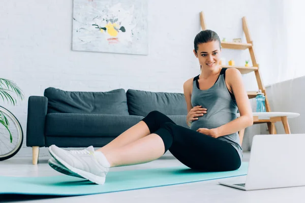 Concentration sélective d'une sportive enceinte regardant un ordinateur portable assis sur un tapis de fitness à la maison — Photo de stock