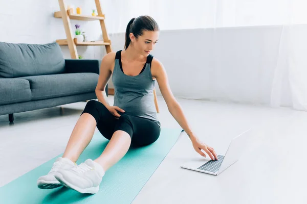 Selective focus of pregnant sportswoman using laptop on fitness mat at home — Stock Photo