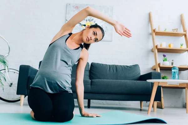 Schwangere Sportlerin streckt Arm, während sie zu Hause auf Fitnessmatte sitzt — Stockfoto
