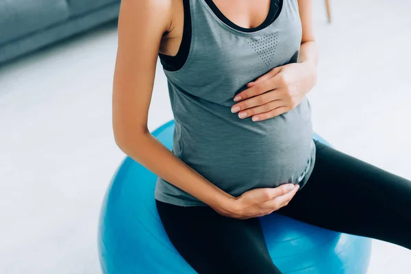 Cropped view of pregnant woman in sportswear touching belly on fitness ball at home — Stock Photo