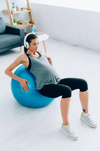 Pregnant sportswoman in headphones training with fitness ball at home — Stock Photo