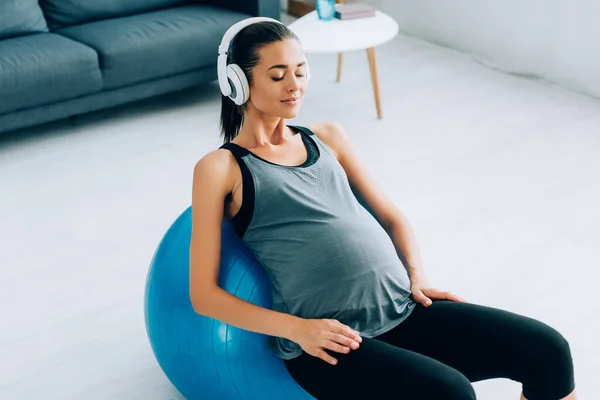 Беременная спортсменка слушает музыку в наушниках, тренируясь дома с фитнес-мячом — стоковое фото
