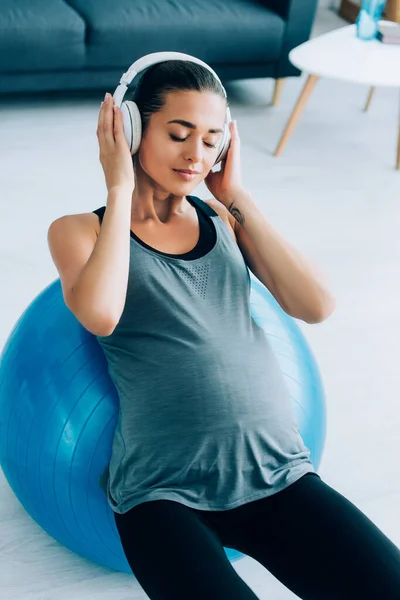Young pregnant woman listening music in headphones while exercising with fitness ball at home — Stock Photo