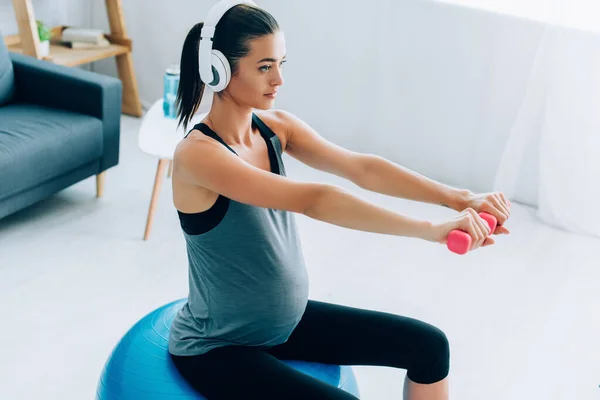 Pregnant sportswoman training with dumbbells and fitness ball while listening music in headphones — Stock Photo