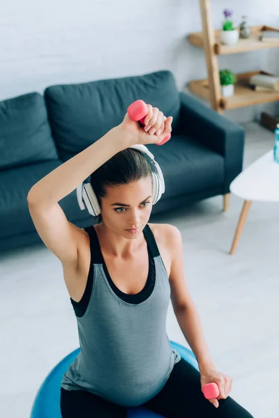 Selective focus of pregnant woman in headphones working out with dumbbells and fitness ball in living room — Stock Photo