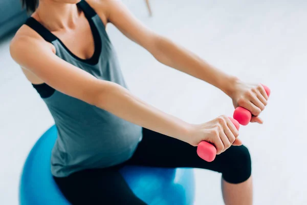 Vista recortada de la mujer embarazada en ropa deportiva haciendo ejercicio con pesas y pelota de fitness - foto de stock