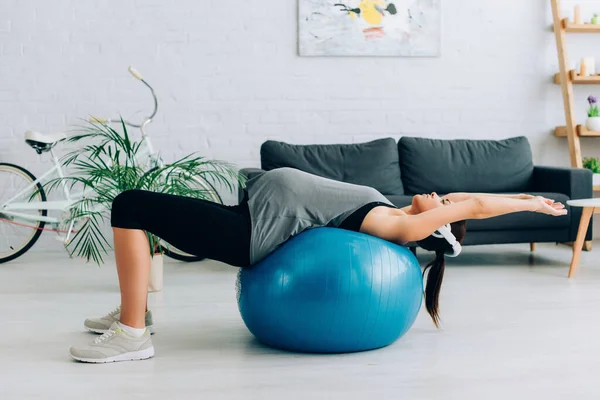 Vista lateral de la deportista embarazada en los auriculares de entrenamiento en la pelota de fitness en la sala de estar — Stock Photo