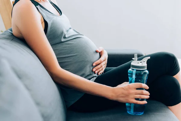 Cropped view of pregnant sportswoman holding sports bottle and touching belly on sofa — Stock Photo