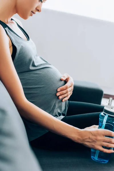 Selective focus of young pregnant sportswoman with hand on tummy holding sports bottle on couch — Stock Photo