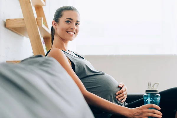 Concentration sélective de la sportive enceinte regardant la caméra et tenant une bouteille de sport sur le canapé — Photo de stock