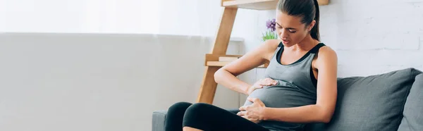 Panoramic shot of pregnant woman in sportswear feeling pain on couch at home — Stock Photo