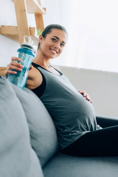 Foco seletivo da mulher grávida em sportswear segurando garrafa de esportes e olhando para a câmera na sala de estar — Fotografia de Stock