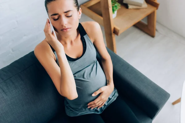 High angle view of pregnant sportswoman talking on smartphone while suffering from pain at home — Stock Photo