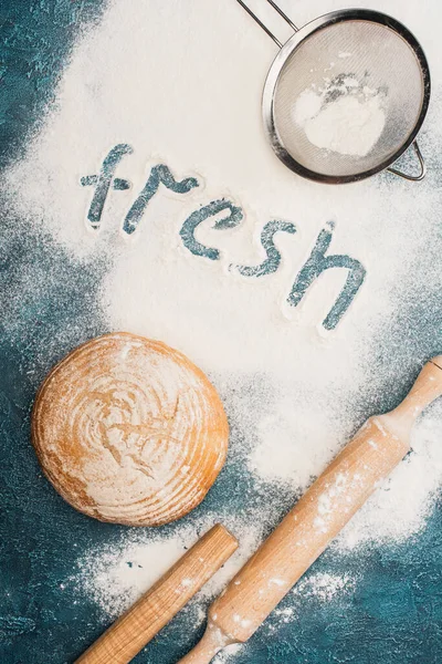 Top view of fresh baked bread loaf near rolling pin, sieve and word fresh on flour — Stock Photo