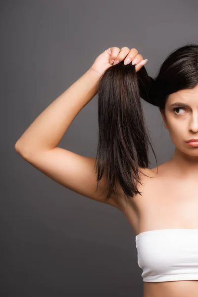 Обрізаний вид засмученої брюнетки жінка тримає пошкоджене волосся ізольовано на чорному — стокове фото