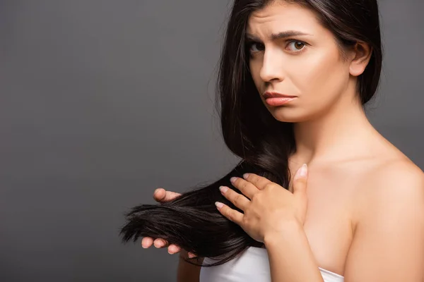 Mujer morena molesta tocando el pelo aislado en negro - foto de stock