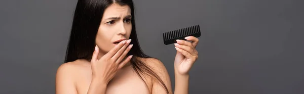 Mujer morena impactada desnuda mirando peine aislado en negro, plano panorámico - foto de stock