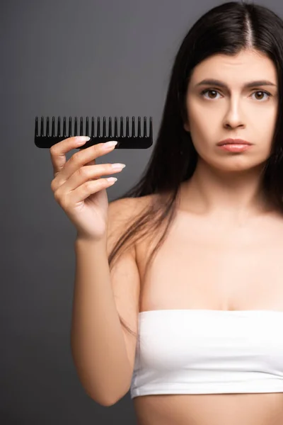 Selective focus of sad brunette woman showing comb isolated on black — Stock Photo