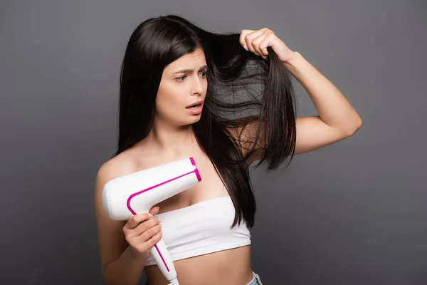 Morena preocupada mujer de pelo largo utilizando secador de pelo aislado en negro - foto de stock