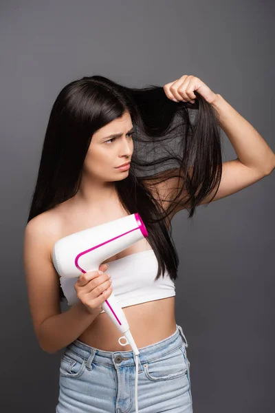 Preocupado morena de cabelos compridos mulher usando secador de cabelo isolado no preto — Fotografia de Stock