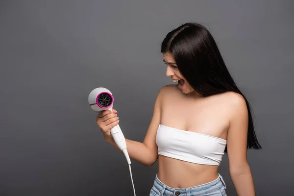 Brunette woman looking at hair dryer and yelling isolated on black — Stock Photo