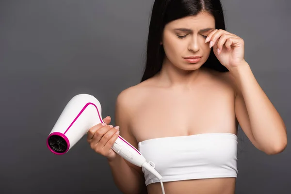 Sad brunette woman holding hair dryer and crying isolated on black — Stock Photo