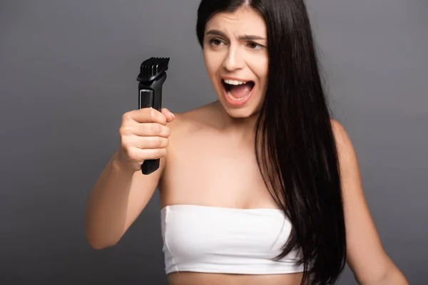 Selective focus of brunette long haired woman holding electric razor and screaming isolated on black — Stock Photo