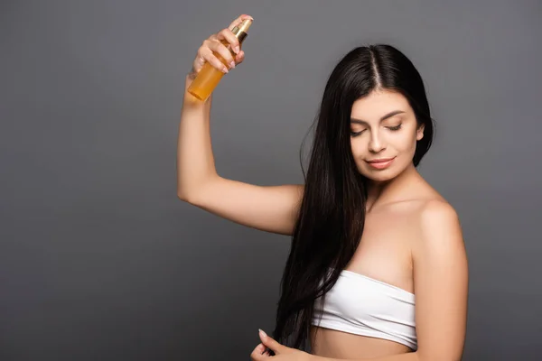 Brunette woman spraying oil on hair isolated on black — Stock Photo