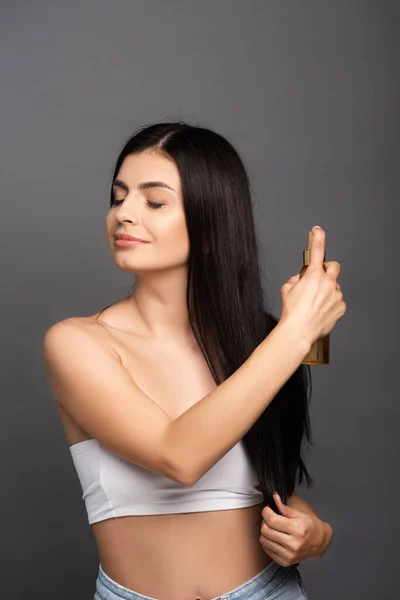 Mulher morena pulverizando óleo no cabelo isolado no preto — Fotografia de Stock