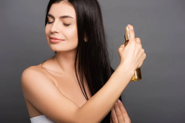 Brunette woman spraying oil on hair isolated on black — Stock Photo