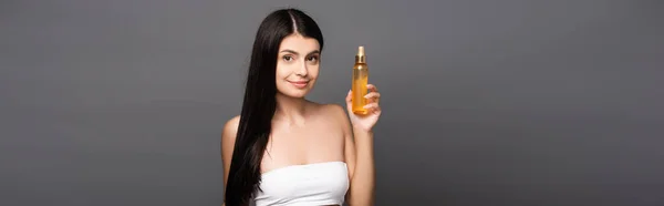Donna bruna con olio per capelli in bottiglia spray e sorridente isolato sul nero, colpo panoramico — Foto stock