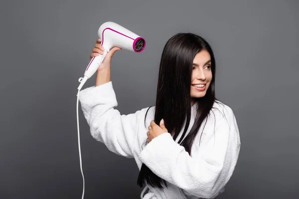 Brunette long haired woman in bathrobe using hairdryer isolated on black — Stock Photo