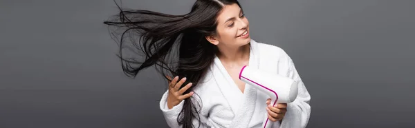 Bruna dai capelli lunghi donna in accappatoio utilizzando asciugacapelli isolato su nero, colpo panoramico — Foto stock