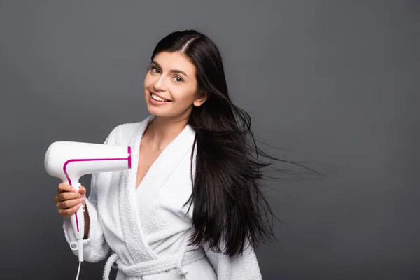 Brunette long haired woman in bathrobe using hairdryer isolated on black — Stock Photo