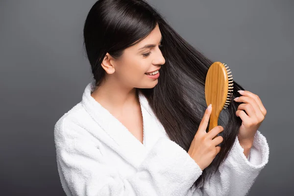 Брюнетка жінка в халаті чистить волосся і посміхається ізольовано на чорному — стокове фото