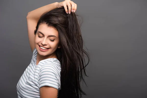 Vista lateral da mulher morena com os olhos fechados sorrindo e tocando o cabelo isolado no preto — Fotografia de Stock