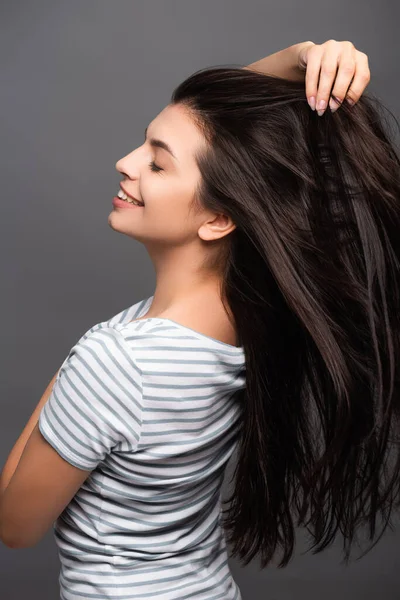 Vista laterale di donna bruna con gli occhi chiusi sorridente e toccante capelli isolati su nero — Foto stock