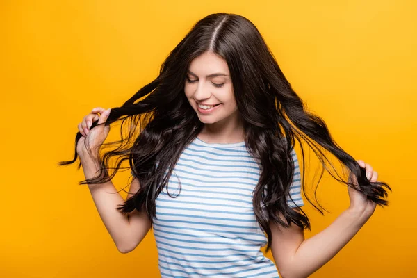 Mulher morena endurecendo cabelos longos encaracolados e sorrindo isolado no amarelo — Fotografia de Stock